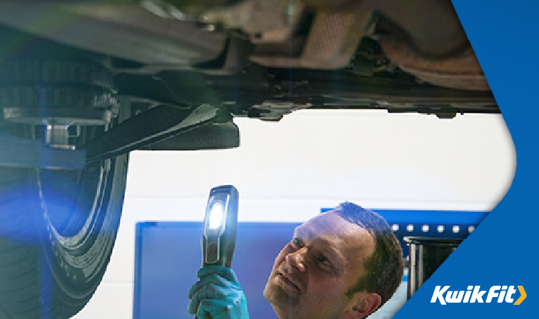 Kwik Fit technician inspecting a tyre with a light underneath a car on a ramp.