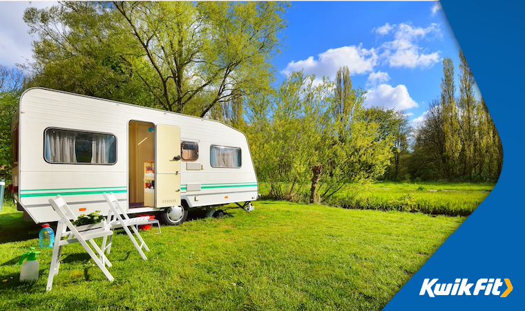 A caravan in a field surrounded by green grass and trees with two chairs pitched up.