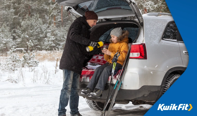 Parent and child at the boot of a car amidst thick snow, warmly wrapped with winter jackets, pouring tea from a thermal flask into a cup.