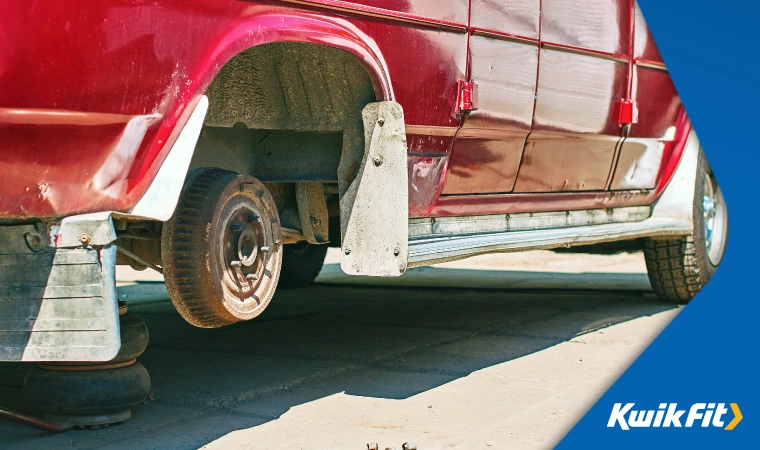 Slightly rusty red classic car raised on jack stands and with its front wheel removed.